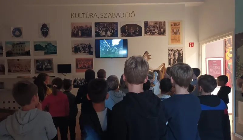Egy múzeumi teremben gyerekek csoportja nézi a falra szerelt képernyőn megjelenő információkat. A képernyő körül régi fotók vannak elhelyezve.