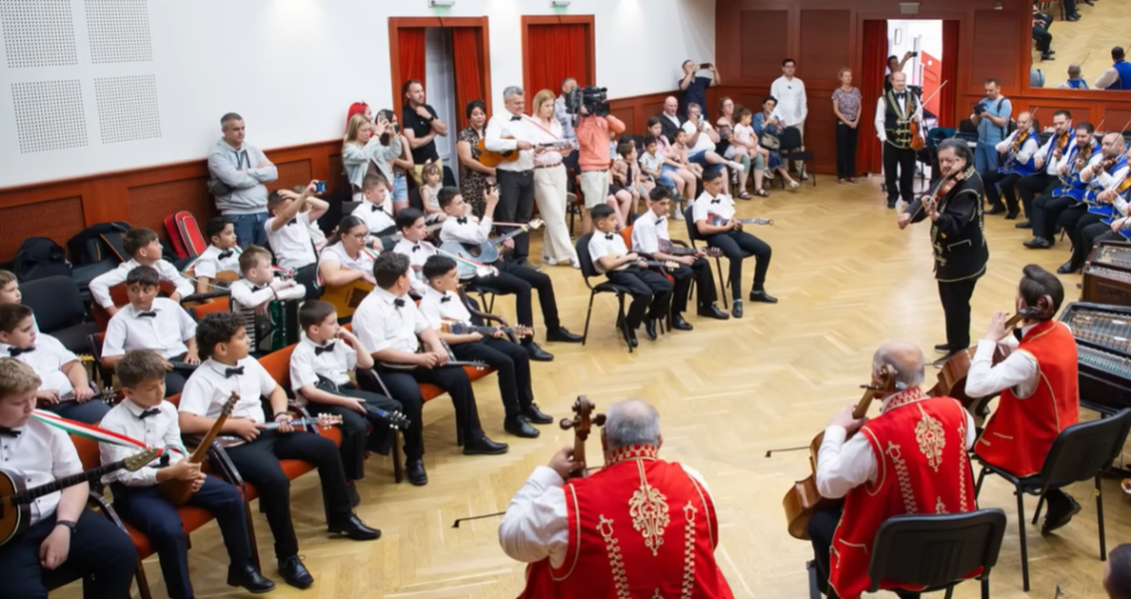 A Csokonai Rendezvényház nagytermében közösen próbál a 100 Tagú Cigányzenekar és a sükösdi SUGO Tamburazenekar. A terem egyik oldalán fehér ingben és nadrágban ülő gyerekek tartják a hangszereiket, velük szemben pedig a cigányzenekar tagjai játszanak piros viseletben nagybőgőn, citerán és hegedűn. Középen fekete ruhában áll a főmuzsikás hegedűvel. A teremben fotósok és videósok rögzítik a pillanatot, miközben néhányan csendben hallgatják a zenét.