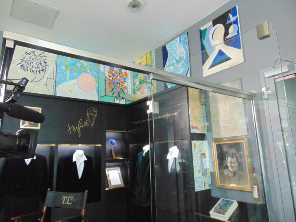 A Tony Curtis Múzeum egyik vitrines kiállítófala látható. A háttérben Tony Curtis portréja és aláírása szerepel, mellette több bekeretezett festmény és grafika, köztük virágmotívumok és absztrakt képek. A vitrin előtt két próbababa áll a művész elegáns, sötét színű öltönyeiben, mellettük egy rendezői szék „T.C.” monogrammal. A kiállítótér megvilágítása kiemeli a relikviákat és a művész személyes tárgyait.