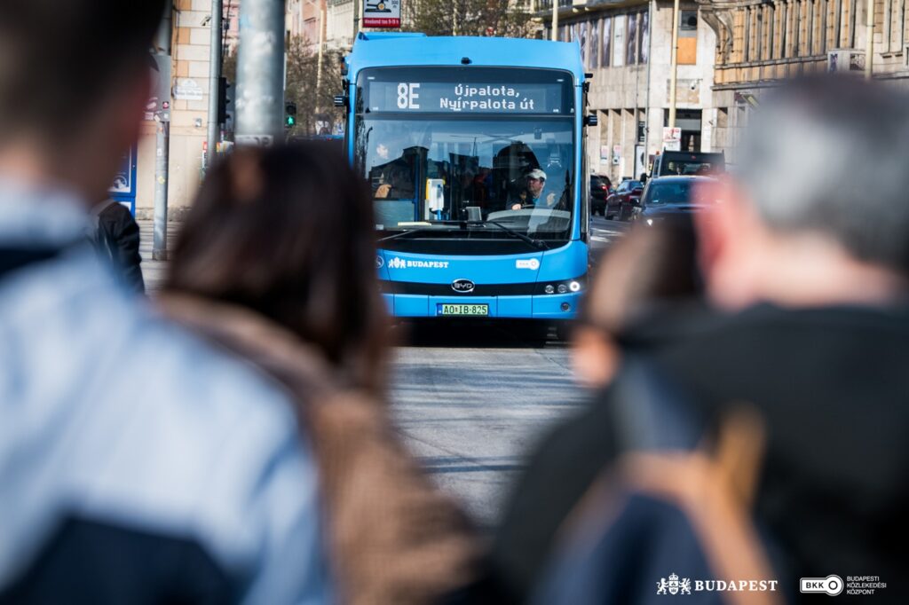 Egy kék, modern elektromos csuklós autóbusz közeledik egy városi kereszteződéshez. A jármű elején a 8E járatszám és az Újpalota, Nyírpalota út felirat látható. A busz előtt néhány gyalogos áll a járdán, mögötte városi épületek és forgalom látszik. A jelenet Újpalotán készült, ahol elindultak az első elektromos csuklós buszok.