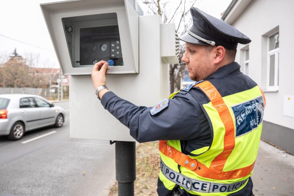 Egy rendőr egyenruhában egy fix traffiboxot állít be az út szélén. A mérőeszköz mögött elmosódva látszanak a nagy sebességgel haladó autók, érzékeltetve a forgalom gyors tempóját.