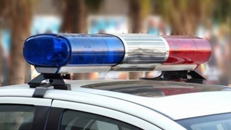 Egy rendőrségi szolgálati jármű tetején elhelyezett kék‑piros villogó lámpa látható közelről. A háttér enyhén elmosódott, fák és kültéri környezet sejlik fel, ami szabadtéri helyszínre utal.