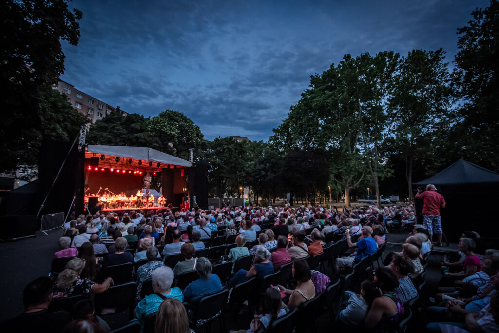 Este, szabadtéri koncert Újpalotán, ahol a 100 Tagú Cigányzenekar lép fel. A nézők nagy számban, szabályos sorokban ülnek a fák között kialakított parkban. A színpadot piros fények világítják meg, a zenekar tagjai hangszereikkel — hegedűk, nagybőgők, cimbalom — a fedett emelvényen játszanak. A háttérben sűrű lombú fák és a nyári alkonyat látszik, a közönség csendben figyel, a koncert bensőséges, ünnepi hangulatot áraszt.