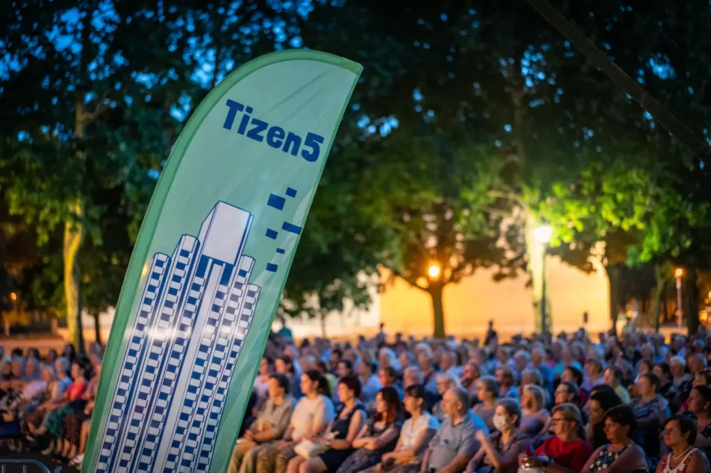 Kültéri esti rendezvény közönséggel, előtérben egy zöld „Tizen5” feliratú zászlóval, rajta stilizált épületgrafikával.