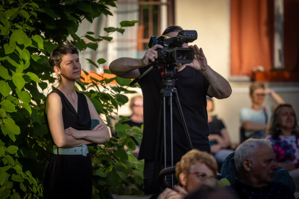 A kép középpontjában szabadtéri rendezvényen egy férfi állványos kamera mögött veszi az eseményeket. Mellette balra egy nő áll, illetve körben közönség ül,. A kép bal oldalán zöld bokor.