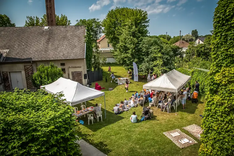 Felülnézetből készített felvétel egy udvarról. A fák közt két függőágy. A füvön két kerti pavilon van felállítva, alattuk székek, asztal. A székeken és előttük pokrócokon az emberek ülnek, figyelik a kicsit hátrébb zajló kutyás bemutatót.